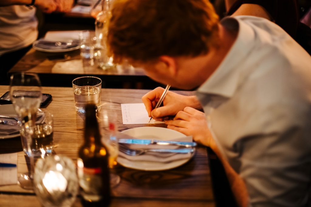 There is a man looking down at a table holding a pen. The man is wearing a white shirt and has a beer next to him.